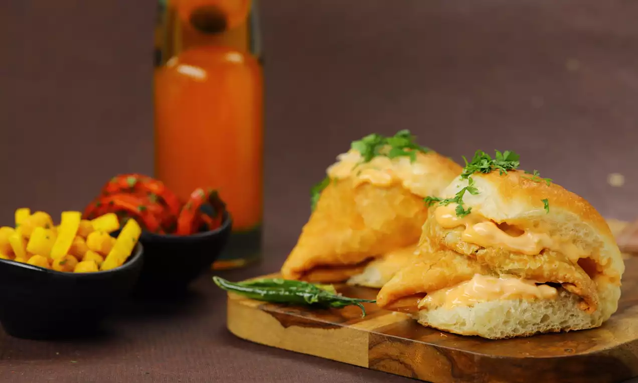 Golden samosas tucked inside soft pav buns with creamy orange sauce, garnished with fresh coriander, served on a wooden board